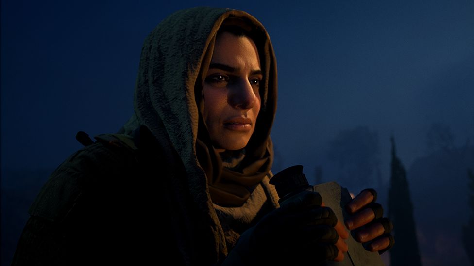 A woman stands against a dark blue night sky, illuminated by the faint glow of a fire. She's wearing a headscarf made of a thick material that covers her hair. She's holding a pair of binoculars at chest level - the lenses are pointed towards the ground. She is concentrating on something in the distance, her expression is impassive.