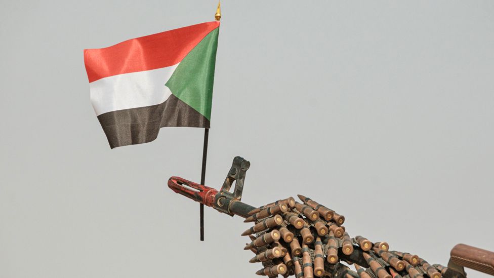 Флаг Судана над дулом пулемета, прикрытым лентой с боеприпасами, военизированных формирований Сил оперативной поддержки (RSF) перед митингом сторонников правящего военного совета Судана в деревне Абрак, 22 июня 2019 г.