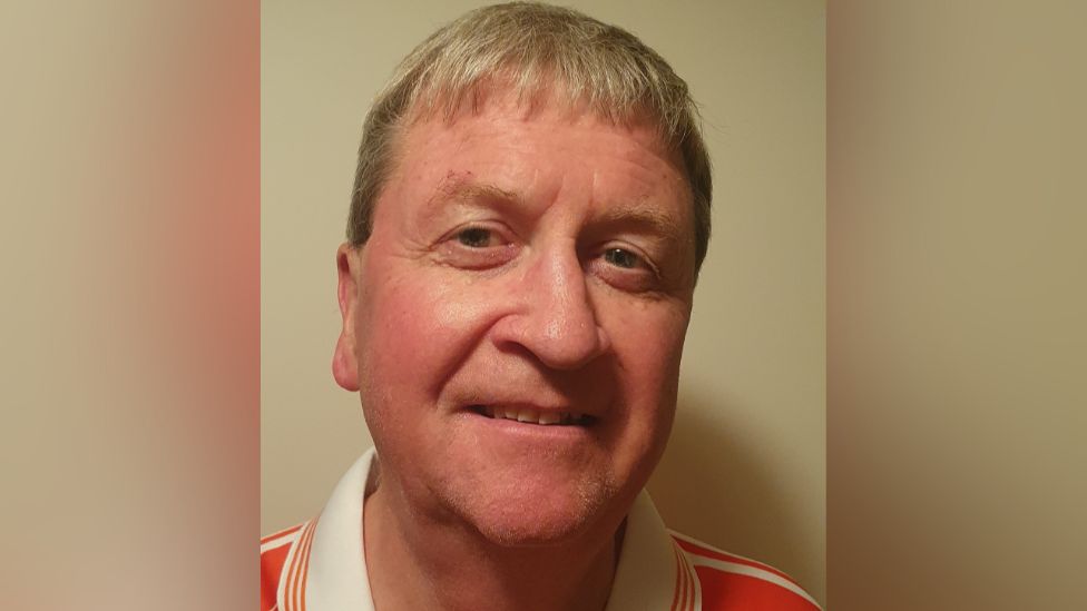 A man with light coloured hair wearing an orange jersey with a white collar smiles at the camera