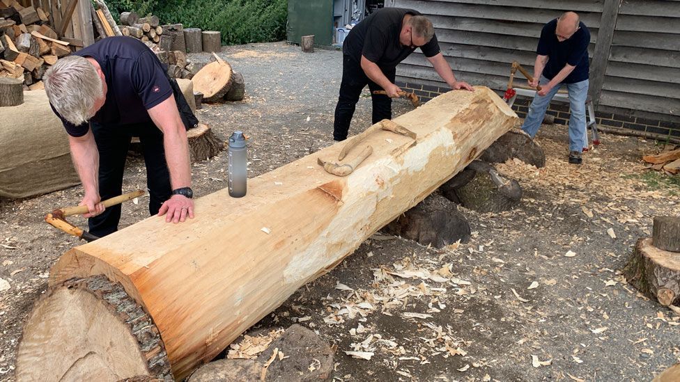 Three men, all wearing black T-shirts, chip away at a large log with tools