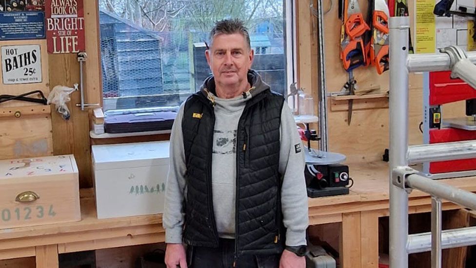 Maintenance manager John Baker in grey hoody and black gilet in front of a repair window at the Emmaus Cambridge workshop