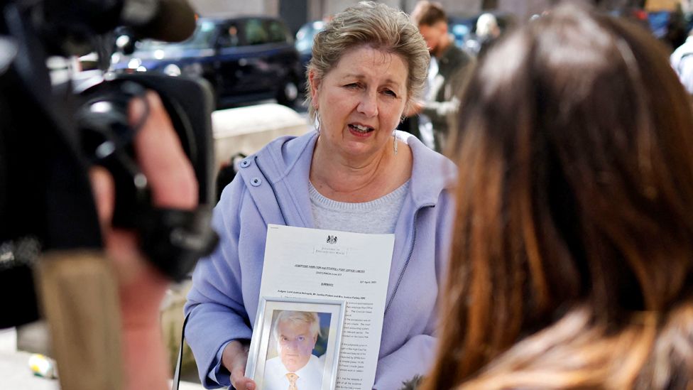 The widow of a former subpostmaster holds his photograph as she speaks to members of the media outside the Royal Courts of Justice in London, on April 23, 2021, following a court ruling clearing subpostmasters of convictions for theft and false accounting