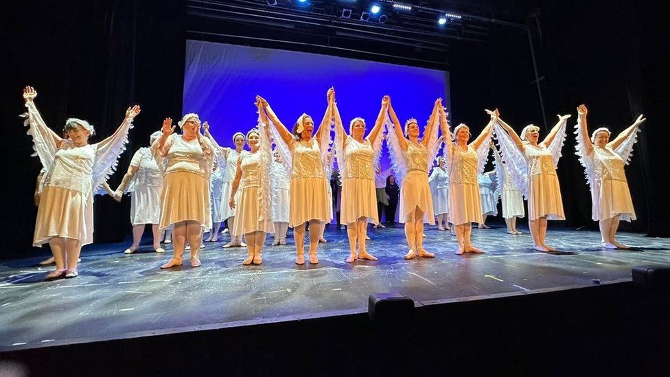 A large group of women in white dresses take a bow on a stage