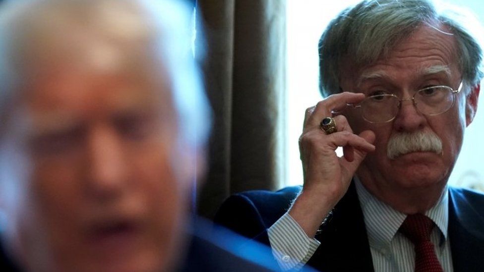 John Bolton adjusts his glasses while looking on at President Trump