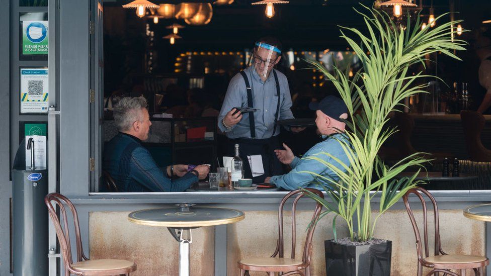 A waiter wearing a face shield takes a payment from customers