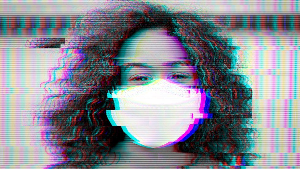 A woman wearing a mask looks at the camera. There is a glitchy effect on the photograph.