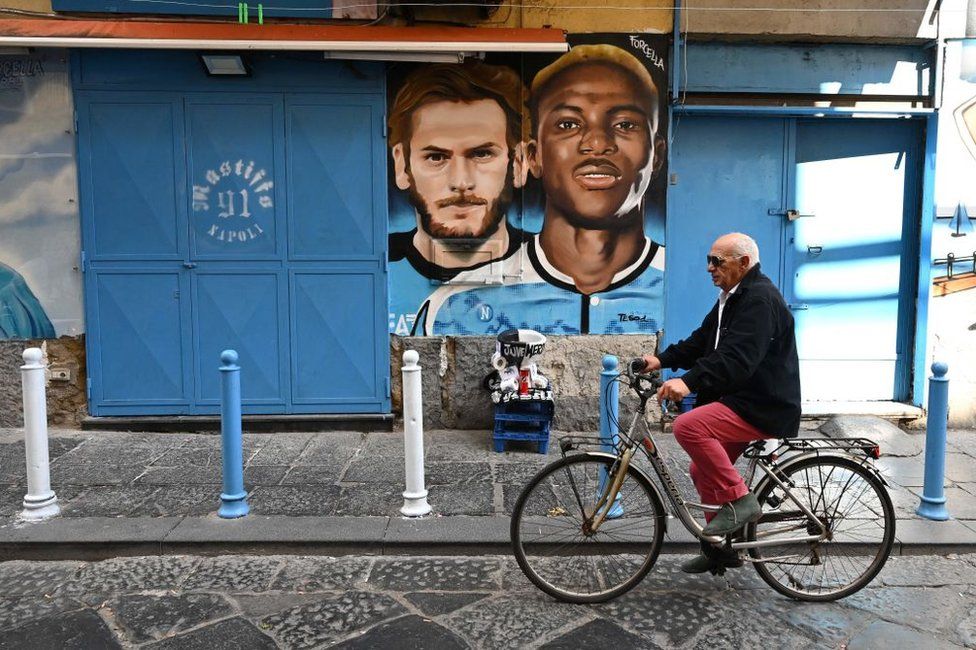A man cycles past a mural in Naples depicting Napoli's Georgian forward Khvicha Kvaratskhelia (L) and Napoli's Nigerian forward , in the Forcella district on March Victor Osimhe, as the city braces up for its potential first Scudetto championship win in 33 years.