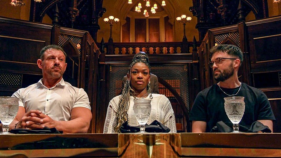 L-R Andrew, Jasmine and Ross at the Round Table in The Traitors. Andrew is a 45-year-old muscular white man with short brown hair. He wears a short-sleeved white shirt with his fingers interlocking in front of him on the table. Jasmine is a 26-year-old black woman with long braids. She wears a long-sleeved crochet cream top. Ross is a 28-year-old white man with short dark brown hair and a short beard. He wears tortoiseshell glasses and a silver chain over a black T-shirt. They are pictured inside the castle with dark wooden carved features and hanging lights. They each have a glass of water in front of them.