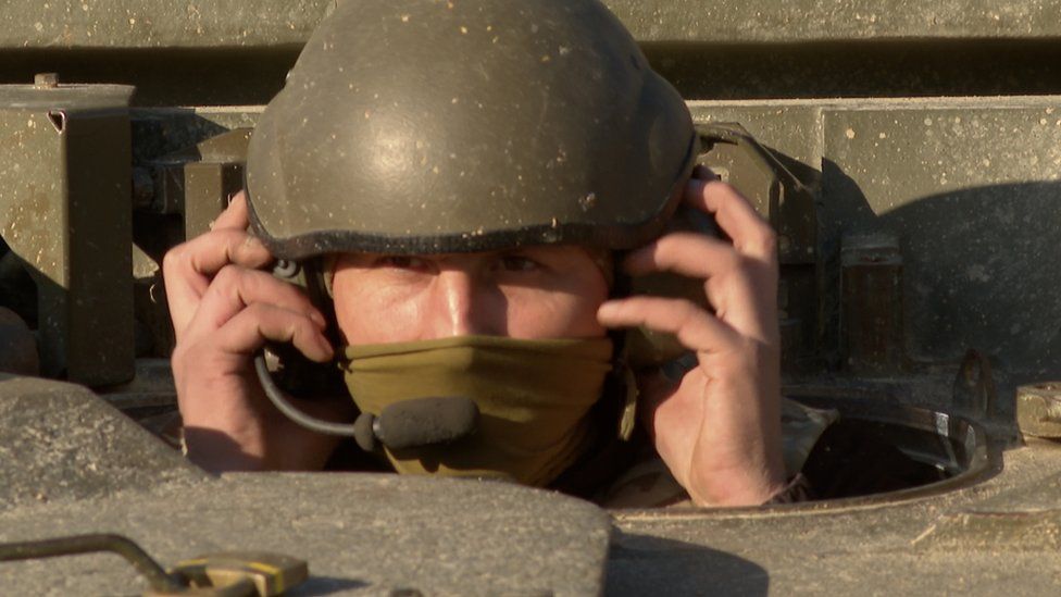 A man hols ear muffs over his ears as he gets out of a tank