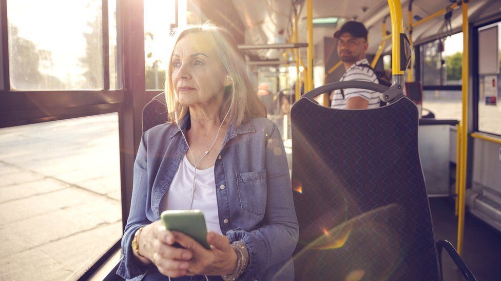 A woman wearing a purple cardigan is listening to her phone through earplugs on a bus.  A man in a cap is looking in her direction.