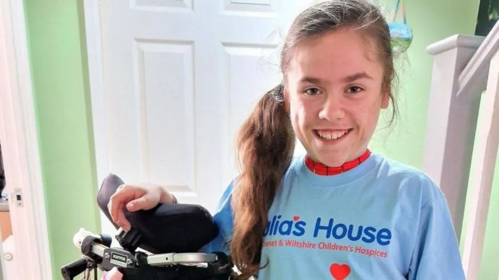 Carmela, from Wareham wearing a light blue julia's house t-shirt over her superhero costume and stood next to her wheelchair