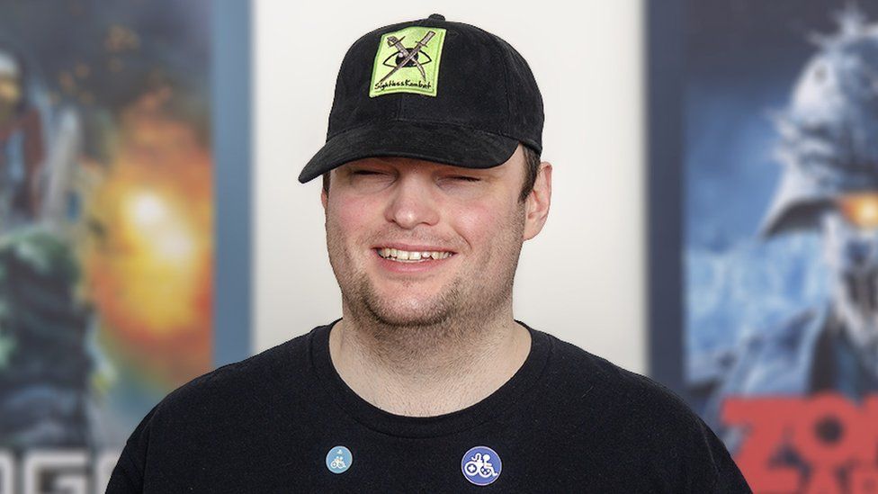 A young man smiling. The logo on his black baseball cap is a lime green square with a pair of crossed swords - a curved, katana-style blade and more elaborate, serrated weapon - over the top of an eyeball graphic. Underneath the words "Sightless Kombat" are written in black script. He's wearing a black t-shirt with an RNIB (Royal National Institute of Blind People) logo on it, and two round pin badges just below the neck line. Both of them have a white, graphic design that mixes a gaming joypad with the seated stick figure more commonly seen with a wheelchair on disabled signage. Behind him two pictures hang on the wall - one shows comic book character Judge Dredd and the other a zombie soldier whose eyes glow yellow underneath his tattered military helmet.