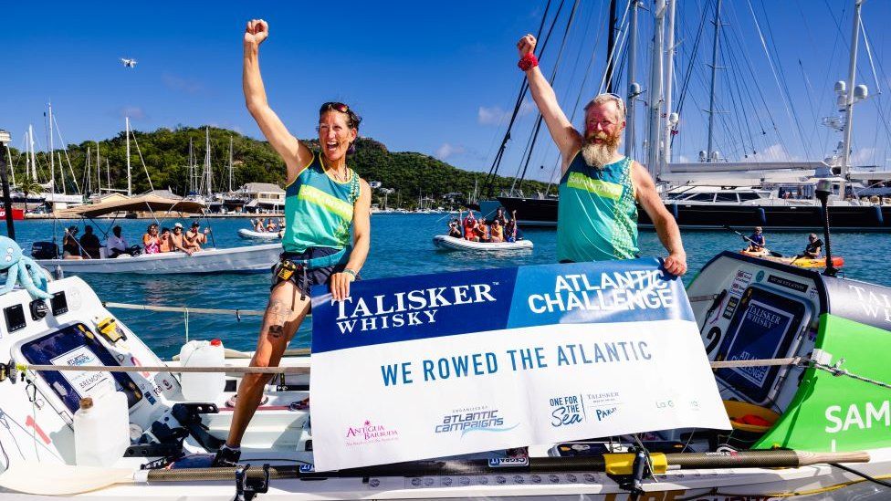 The couple stand, one arm punching the air each, standing with a sign saying they completed the Atlantic challenge