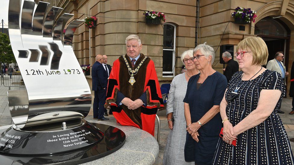 The mayor and relatives of victims look at the new memorial, a metal structure on top of a plinth