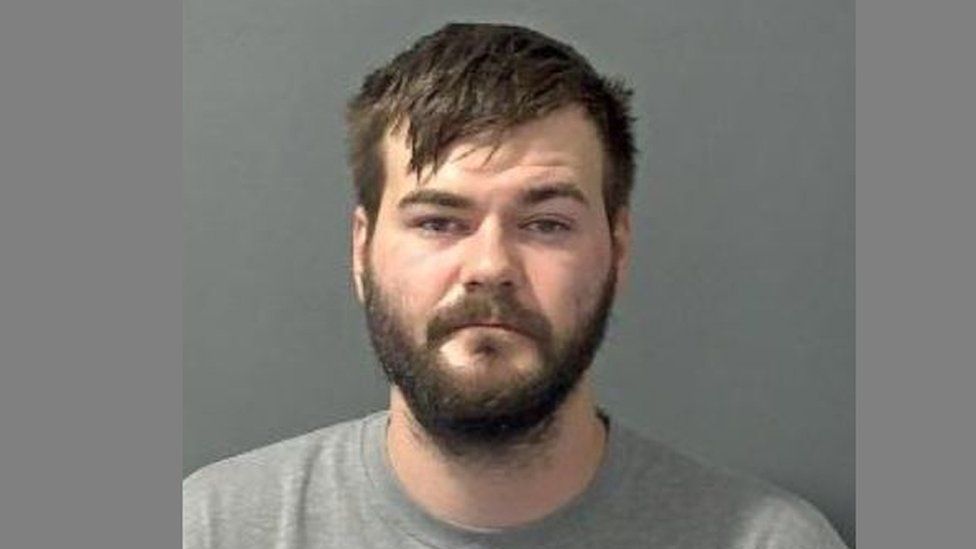 Police "mug shot" of a man with a dark beard wearing a grey t-shirt