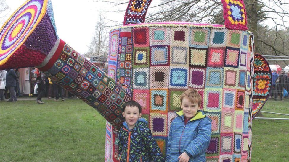 Two boys standing in front of a giant knitted teapot