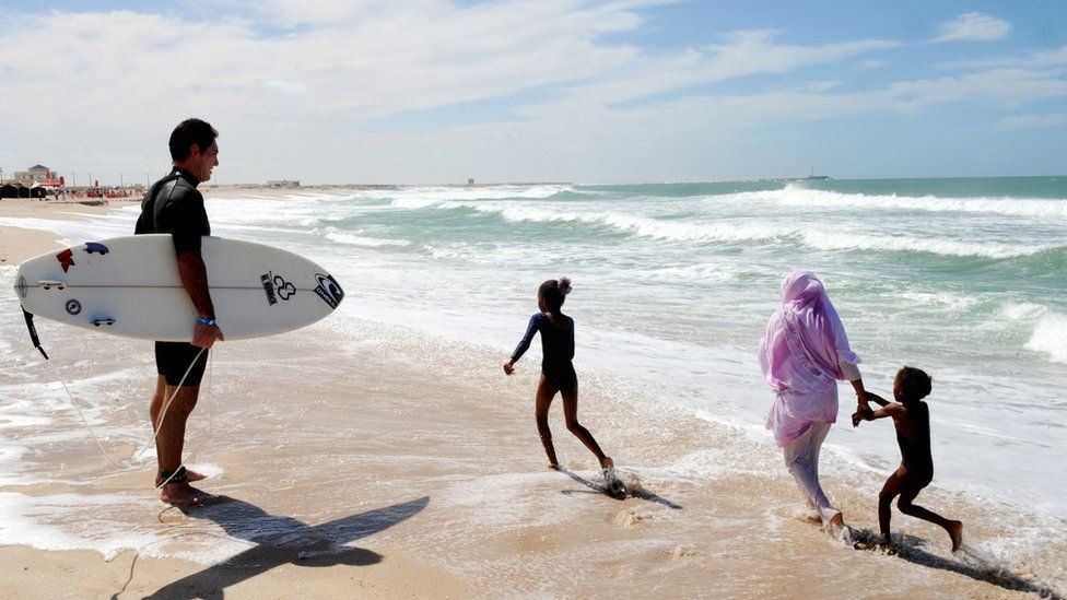 A surfer watches a Western Saharan mother and children run along the beach.
