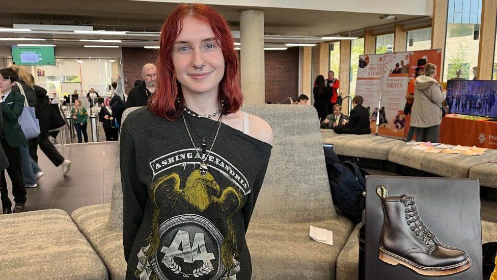 Woman with red hair and green t-shirt poses in exhibition hall with Doc Martens Shoe