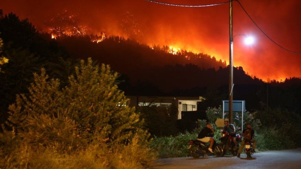 People gather on motorbikes as wildfire flames rise behind them on Evia