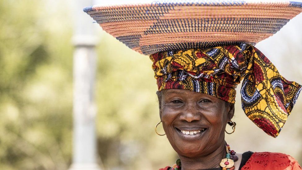 Woman wearing tradition clothes with a basket on her head. She is wearing a headwrap and is smiling.