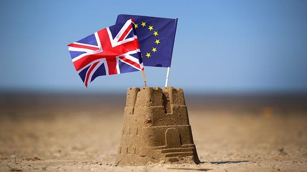 The flag of the European Union and the Union flag sit on top of a sand castle on a beach