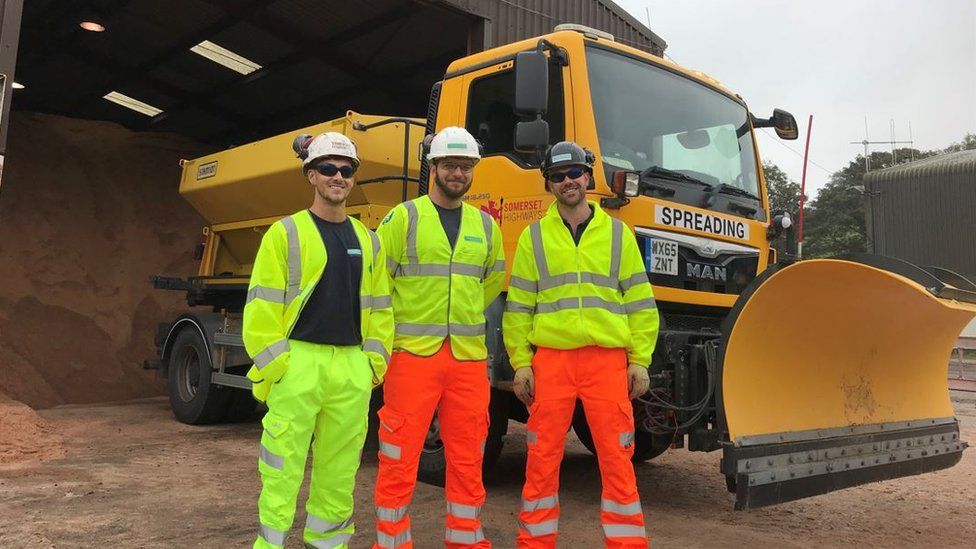 Josh, Jordan And Rich From Somerset Council's Gritting Team wearing hi-vis jackets and hard hats, standing in front of a yellow gritting truck with a plough on the front