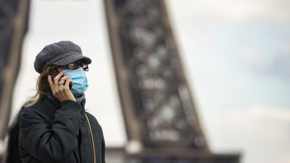 A woman wearing a surgical face mask walks near the Eiffel Tower