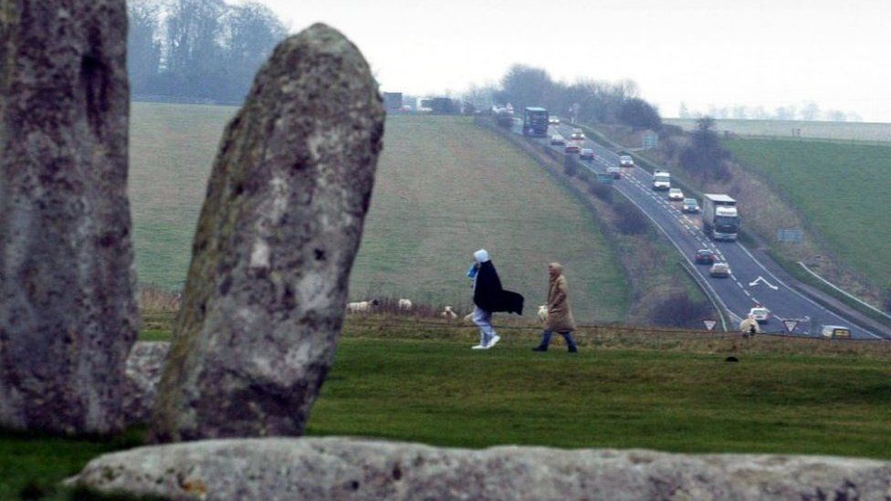 People walking past Stonehenge as cars drive down the A303 in the distance