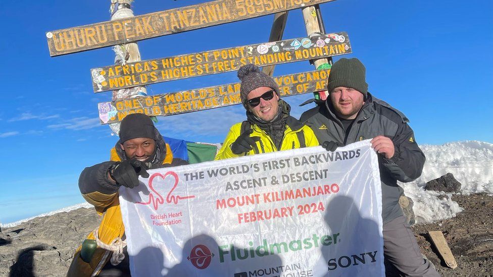 Three men at the top of a mountain with a 'World's first backwards ascent' sign