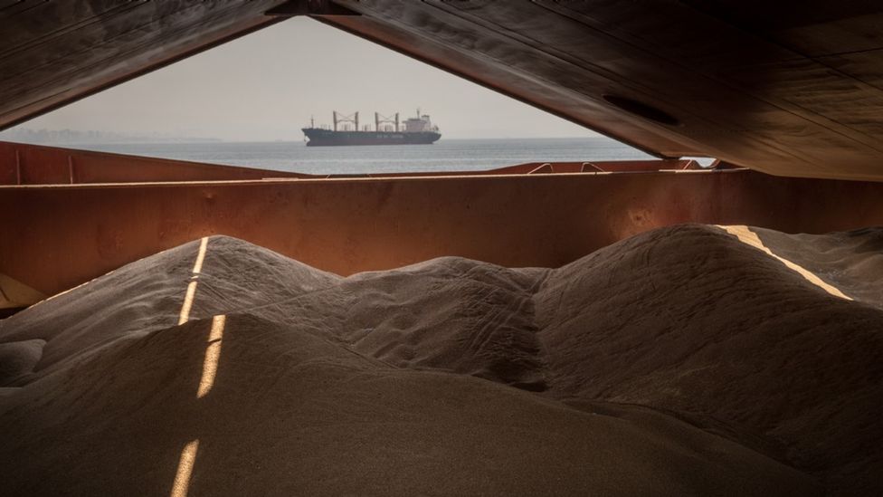 View of a cargo ship at sea, carrying grain from Ukraine, seen through the opening of a hold of another ship, with piles of grain in foreground.