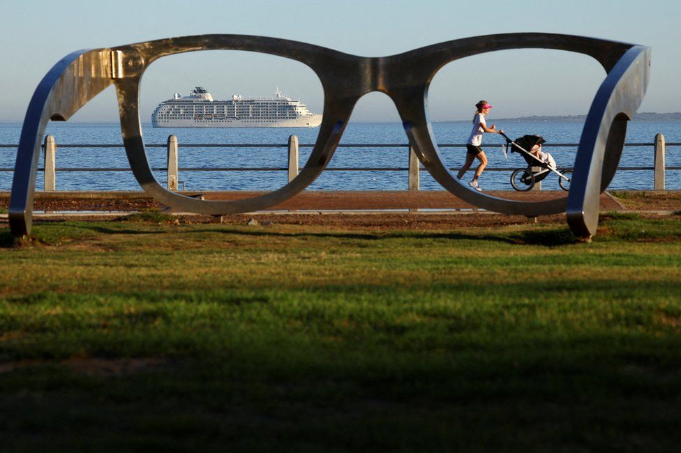 A woman runs with a pram in the foreground, and a private residential ship is seen in the background. Both are framed by a sculpture in the shape of reading glasses.