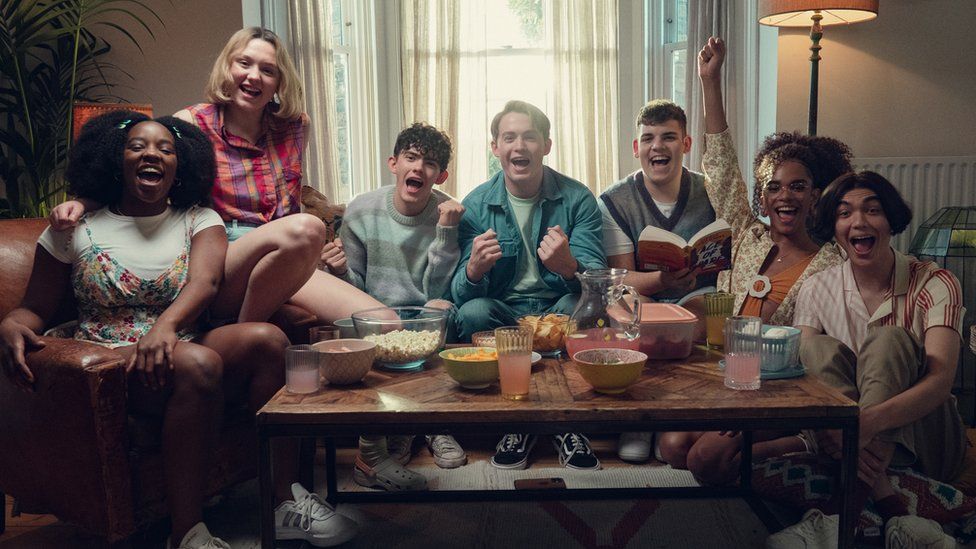 Seven teenagers sit on and around a sofa in a living room. There's a coffee table in front of them packed with bowls of snacks and pitchers of juice. There's a young, black girl on the far left with her hair in bunches, she's laughing. A young, white girl wearing a sleeveless, pink and purple striped shirt and blue shorts. sits next to her, her arm round her shoulder. Next to her is a young, white man with dark curly hair. He's wearing a stripy jumper and has his fists clenched in a celebration pose. Next to him is another young white man with curtains haircut, pulling a similar pose. Next to him is a young white man with dark hair wearing a v-neck tank top and holding a book. Next to him is a young black female wearing a floral shirt and holding one arm in the air with fist clenched, but smiling widely. Finally, one young man, mouth open wide as if saying "hurray", with a stripy shirt and dark, curtain-style hair, sits on the floor at the end of the row.