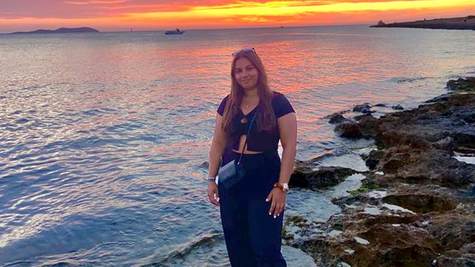 Shayla Rathod stood smiling for the camera on a rocky beach right by the water's edge. She is has sunglasses on the top of her head and her dark hair is worn down over her shoulders. She is wearing a navy top and navy trousers and has a black handbag over her shoulder hanging in front of her midriff. She has a watch on her left wrist. Behind her is the sea and a pretty orange sunset with a boat also visible in the background