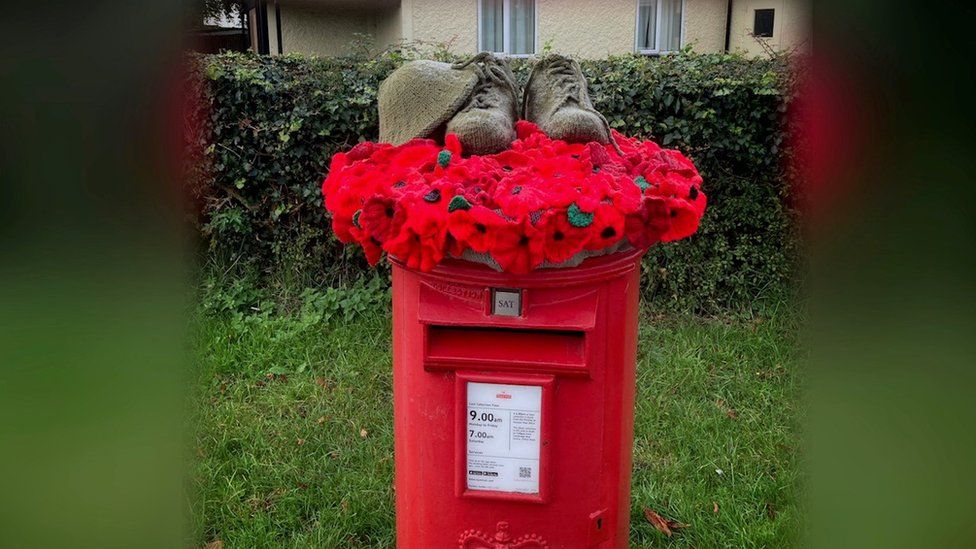 Red postbox on grass with a hedge behind. A garland of poppies lies on top, with knitted soldier's shoes and helmet