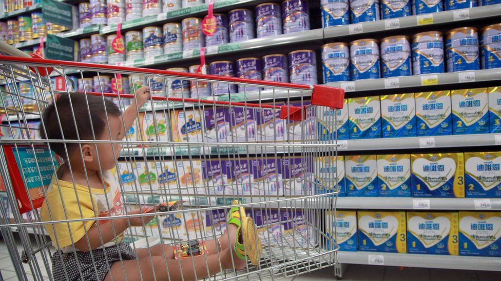 Child sits in a shopping trolley in a Chinese supermarket, with tins of milk formula on shelves in the background