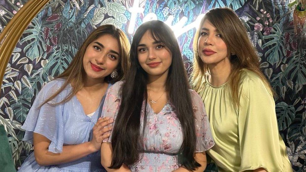 Aliza Ayaz (left) with her sister and mother. Aliza is a 25-year-old South Asian woman with long brown hair worn loose. She wears a sparkly blue blouse and leans into her sister, who also has long dark hair and wears a light pink top. Her mother is on the end and wears a pale green top. They are pictured in front of a wallpaper decorated with leaves and foliage.