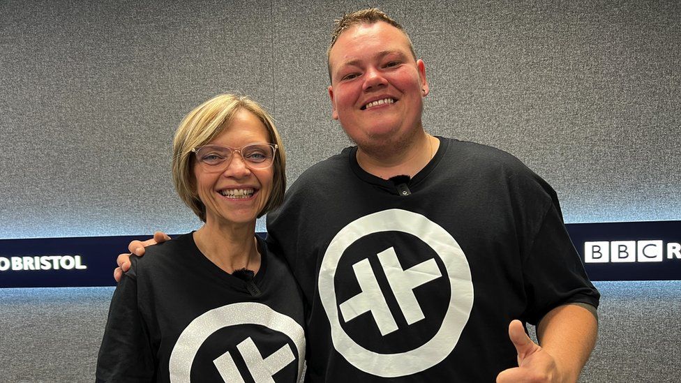 Denise Coles and Joe Sheppard. They are both wearing black T-shirts printed with a white Take That logo - two rotated Ts inside a circle. Joe has his arm around Denise and both of them are smiling at the camera