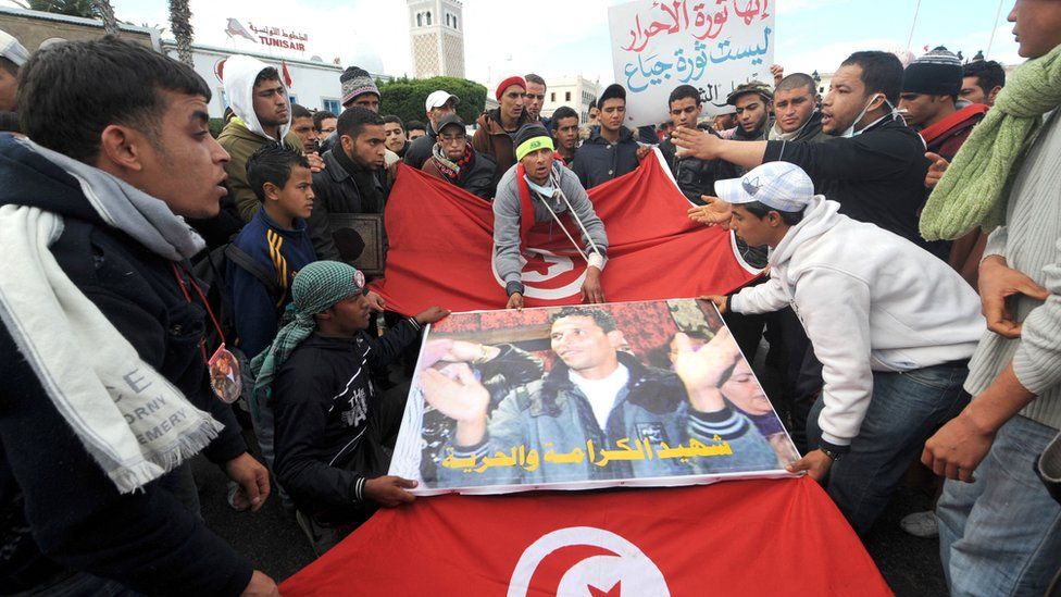 People walk with Mohamed Bouazizi poster and Tunisian flag in front of the government palace in Tunis on January 28, 2011.