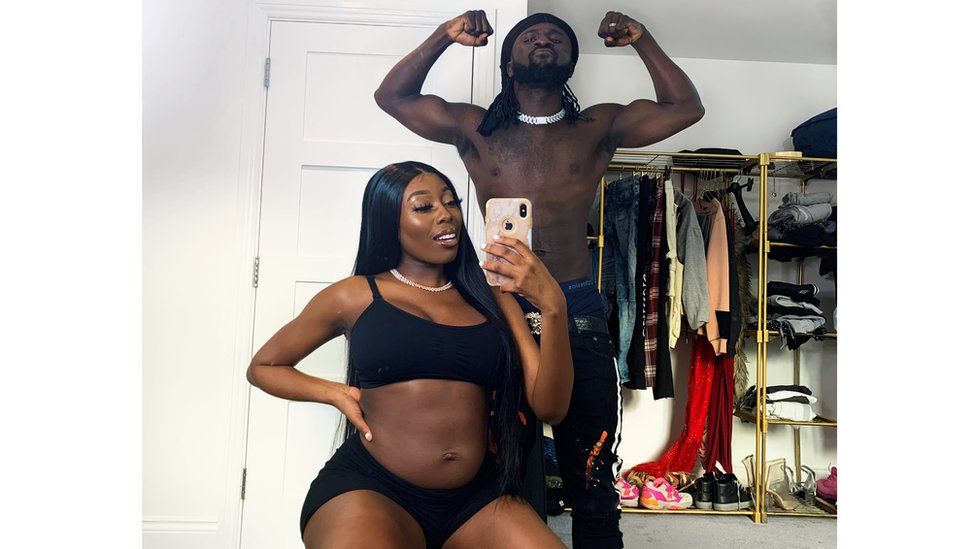 Global Boga and Nicole Thea posing together in a mirror seflie photograph. Nicole is holding the phone in her left hand with her right hand on her belly. Global is doing a muscle pose. The background has a white door which is closed, and a golden clothing rail in the back with an assortment of clothes hanging on silver wire hangers.