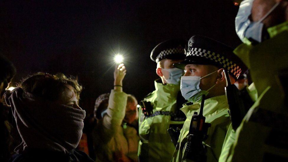 A shot taken at close distance of a woman wearing a face masks facing a line of Met police officers, who are all wearing face masks. The photo is taken at night, and in the background someone is holding up a light on their phone.