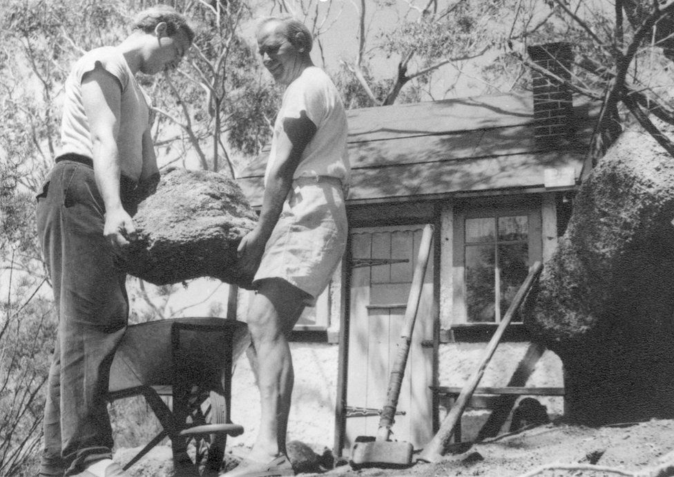 In a black-and-white image, Helmut and Peter Mayer carry a mound of dirt to a wheelbarrow
