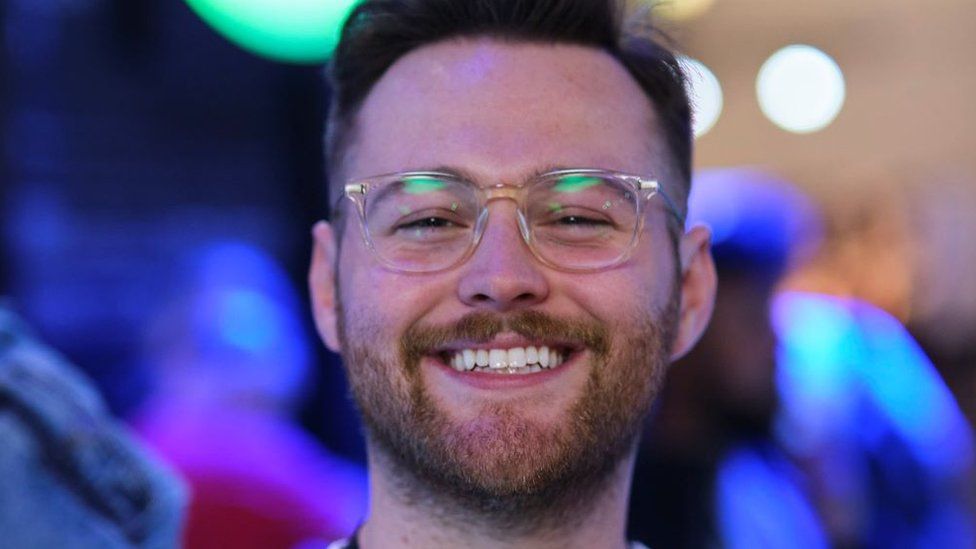 James Hodges. James is a young white man with short brown hair and a trimmed brown beard. He has brown eyes and wears clear-framed square shaped glasses. He is smiling at the camera and is photographed inside at an event - behind him is a blurred background lit by blue lights.