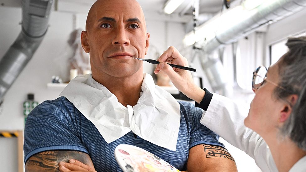 An artist at the Grevin Museum paints the waxwork model of Dwayne "The Rock" Johnson. The artist is a white woman with short grey hair down to her chin. She wears dark rimmed glasses and a white shirt and poses holding a small paint brush up to the waxwork's face. The waxwork depicts a bald Dwayne Johnson and is dressed in a light blue polo shirt. It is modelled with folded arms painted with swirling tribal patterned tattoos showing off the wrestler's biceps. The waxwork looks lifelike and the skin tone has been darkened after earlier being criticised for being incorrect.