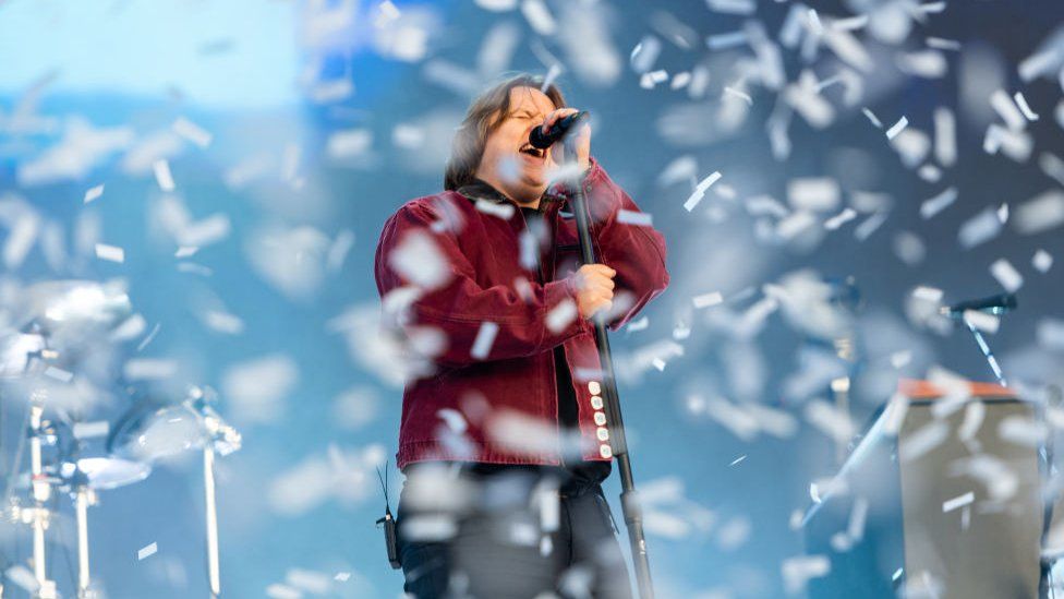 Lewis Capaldi singing on stage at BBC Radio 1's Big Weekend. He is wearing a red jacket and there is a lot of confetti all over the stage in front of him and around him.
