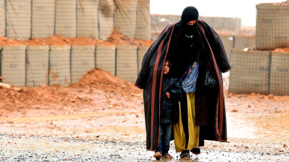 File photo: A Syrian refugee from the informal Rukban camp walks in the rain as she shelters a young child outside a UN-operated medical clinic, 1 March 2017
