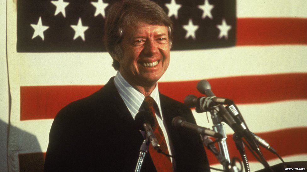 U.S. president Jimmy Carter smiling at a podium in front of an American flag, 1970s.