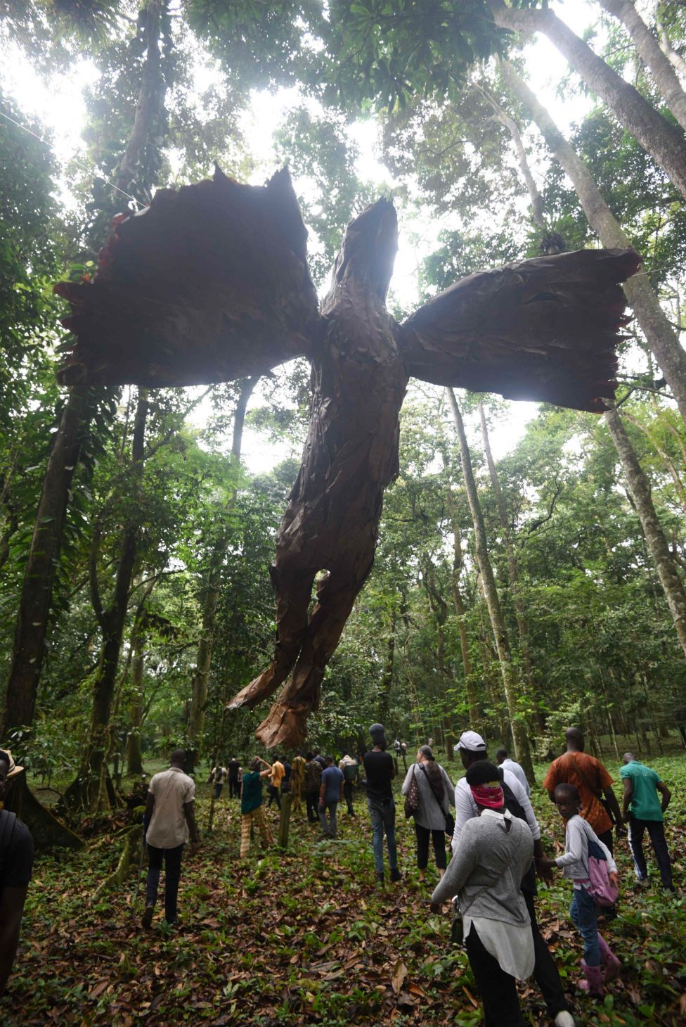 People in a forest walk past a large statue hovering overhead.