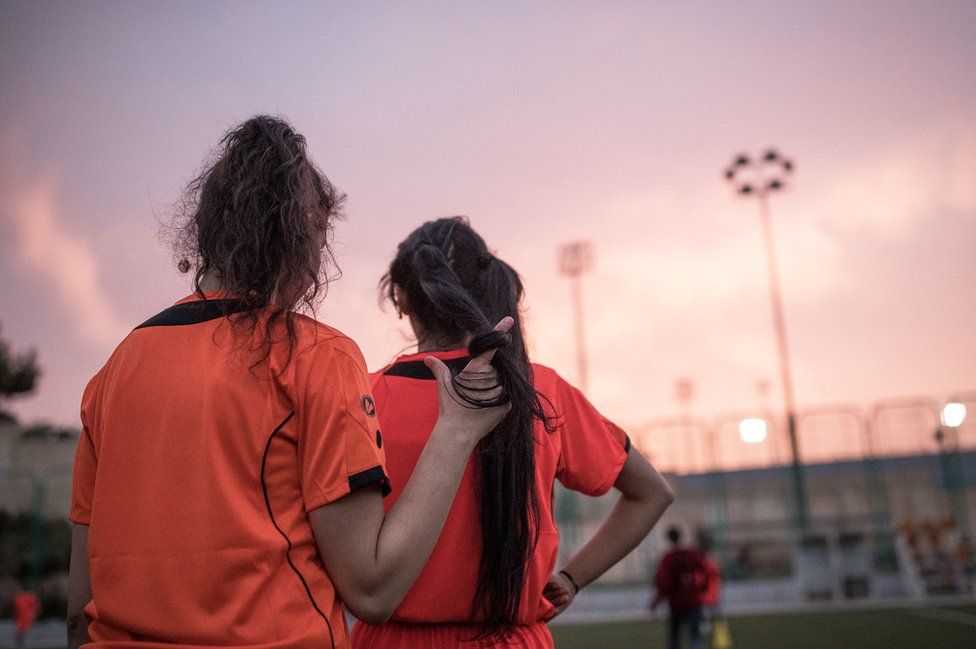 Two girls watch their training session from the sidelines.