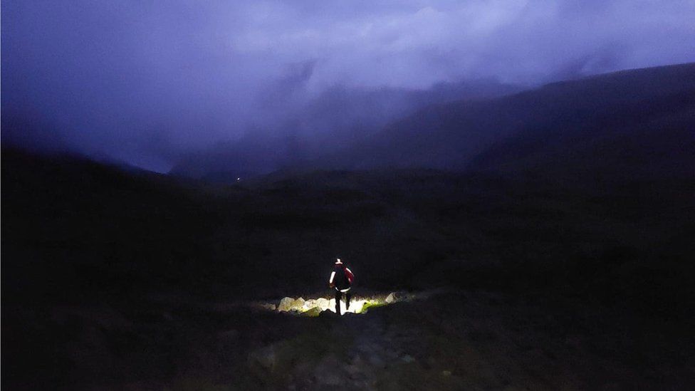 A volunteer illuminated by a head torch walks through dark and misty night