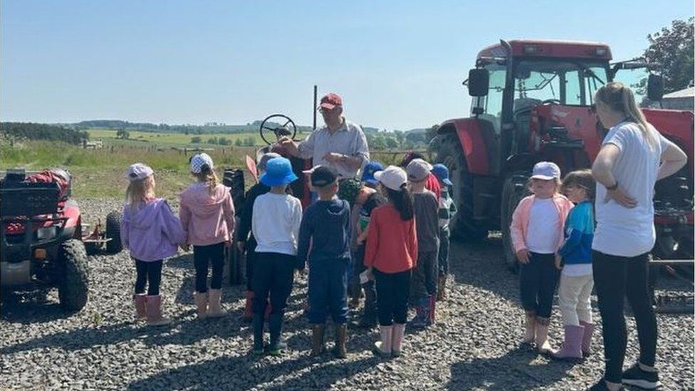 A group of children gather round Charlie Bennett with a teacher watching and a tractor in the background